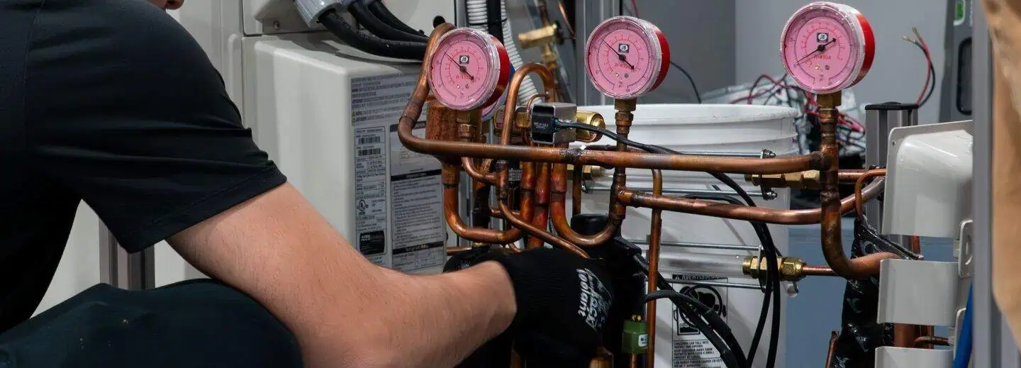 A technician in black work attire adjusts HVAC equipment, focusing intently. Copper piping and pressure gauges are visible, suggesting maintenance work.