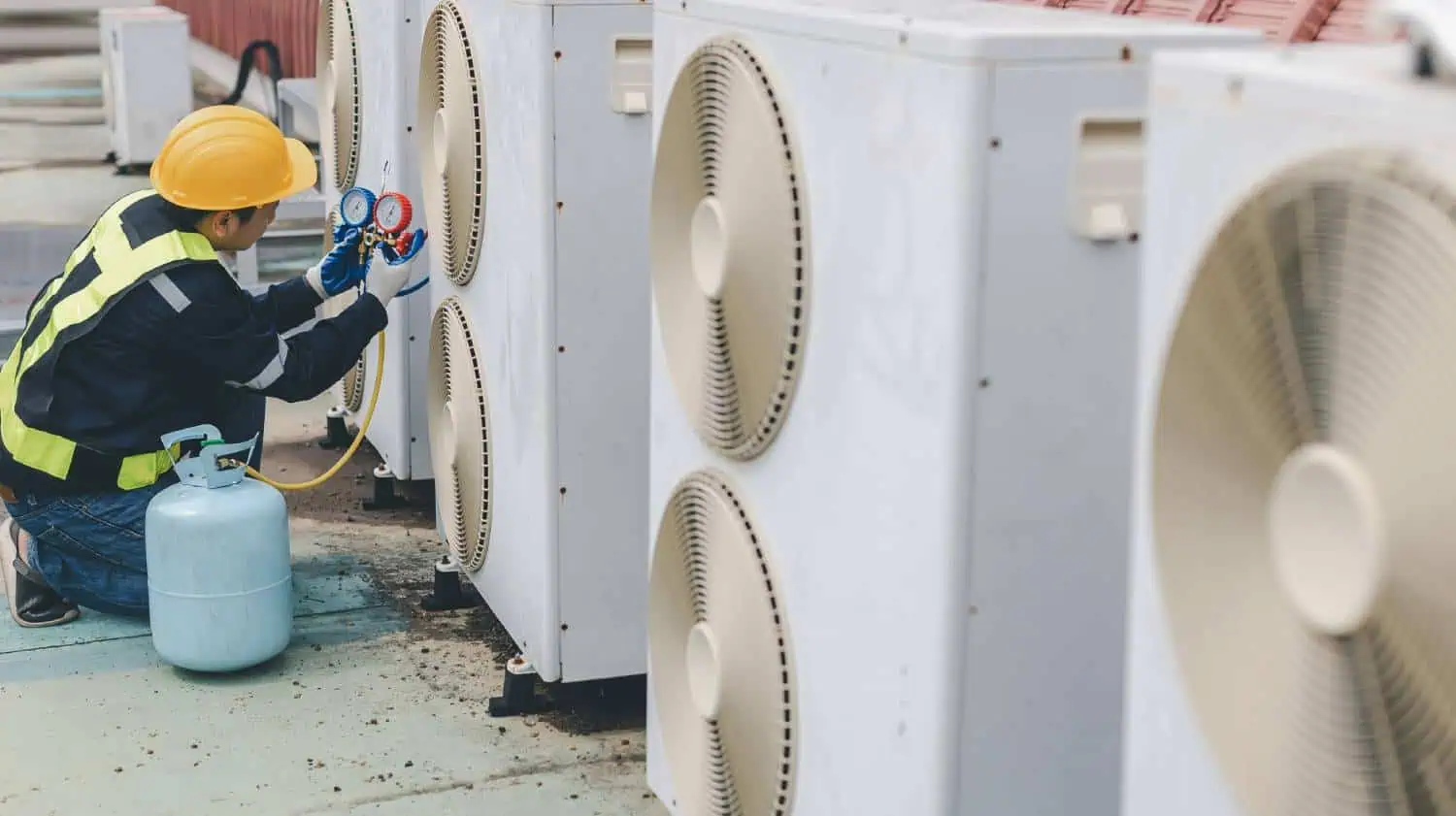 Technician in yellow hard hat checking AC unit pressure with gauges and hoses